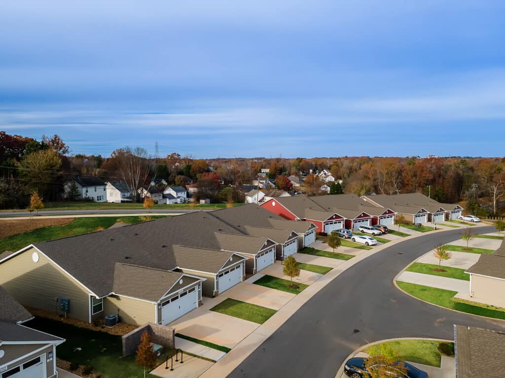 Aerial view of a suburban neighborhood with single-story houses, attached garages, driveways, and a curved street, surrounded by trees with autumn foliage.