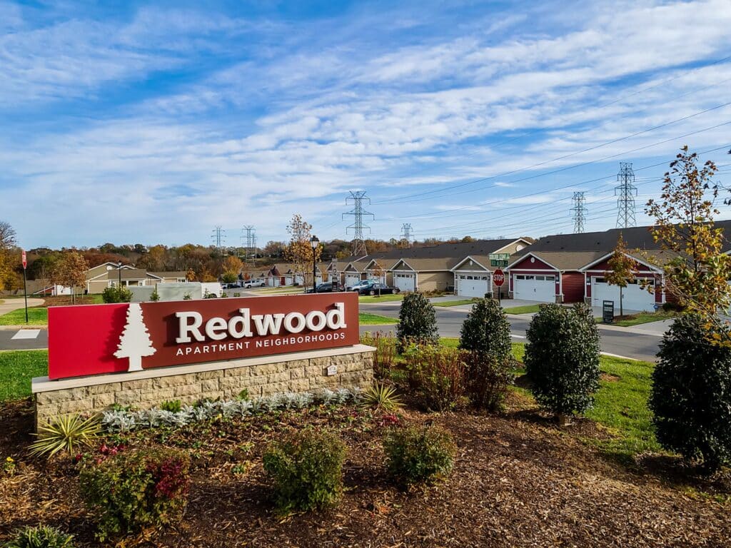 Redwood Apartment Neighborhoods sign in front of a residential area with houses, power lines, and trees under a partly cloudy sky.