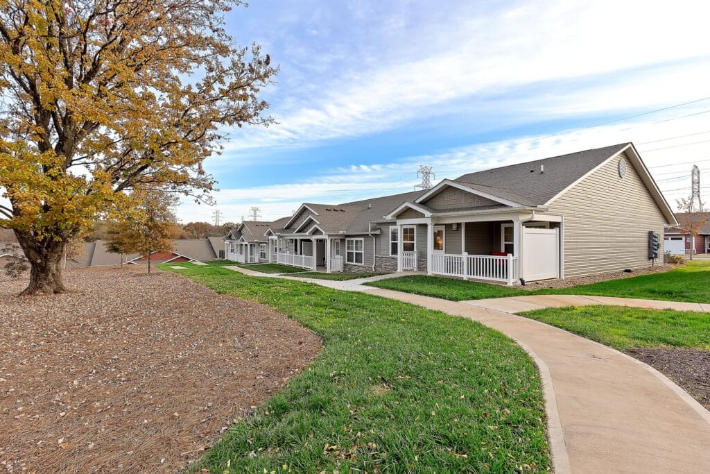 Single-story residential buildings with front porches line a curved sidewalk, bordered by grass and a large tree with autumn leaves.