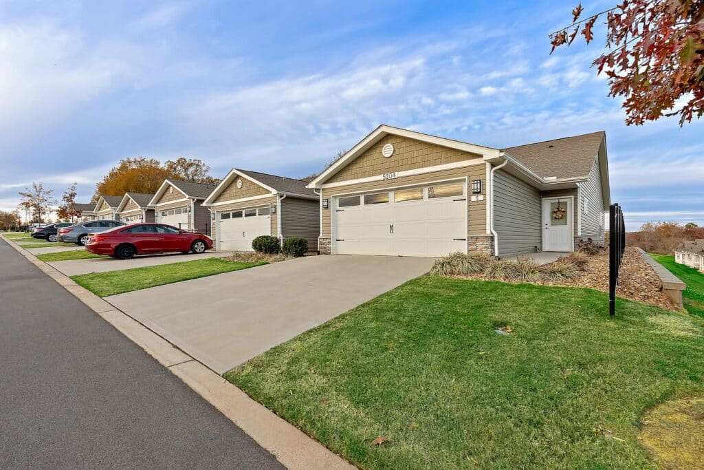 Row of single-story suburban houses with driveways and garages, green lawns, and cars parked along the street under a partly cloudy sky.
