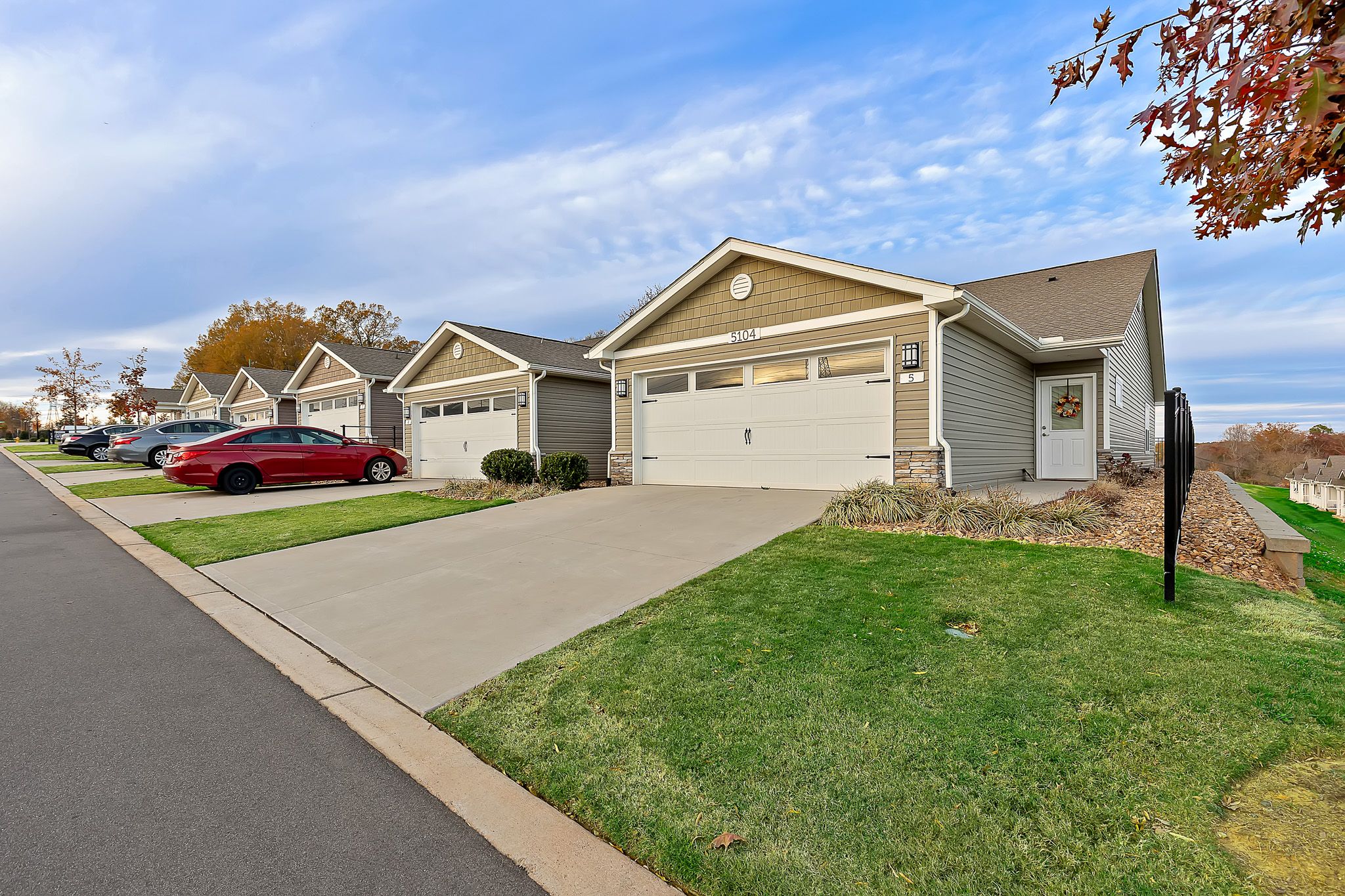 Row of single-story suburban houses with driveways and garages, green lawns, and cars parked along the street under a partly cloudy sky.