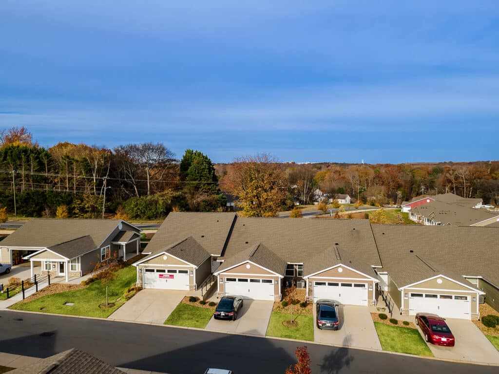 A row of suburban single-story houses with garages, parked cars in driveways, and trees with autumn foliage in the background under a clear blue sky.