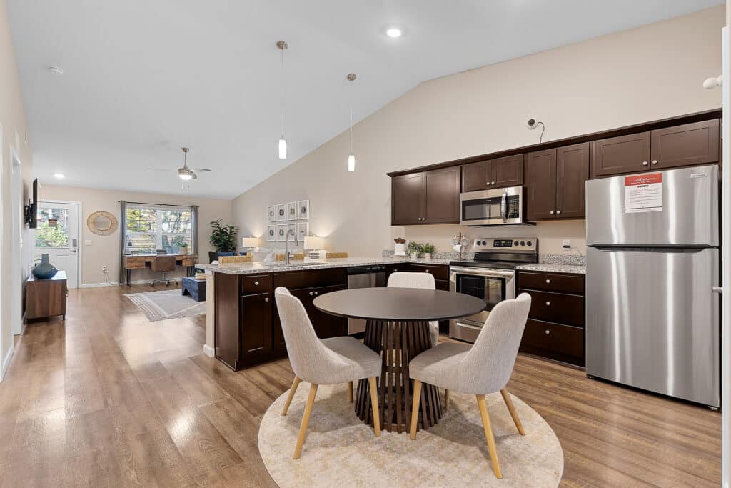 Open-concept kitchen and living area with dark wood cabinets, stainless steel appliances, a round dining table with four chairs, and light wood flooring.