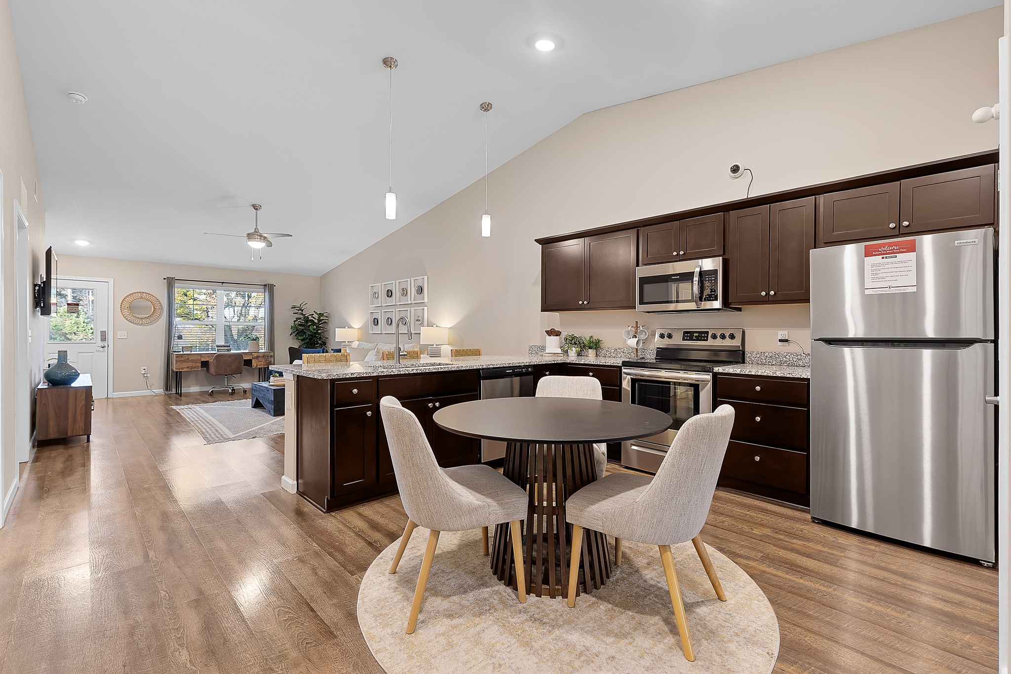 Open-concept kitchen and living area with dark wood cabinets, stainless steel appliances, a round dining table with four chairs, and light wood flooring.