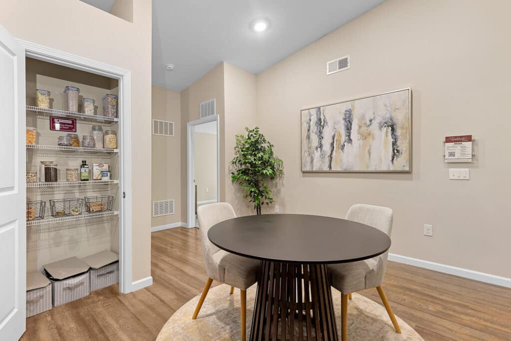 A modern dining area with a round table and two chairs, abstract wall art, a plant, and an open pantry with organized shelves of food items and storage bins.