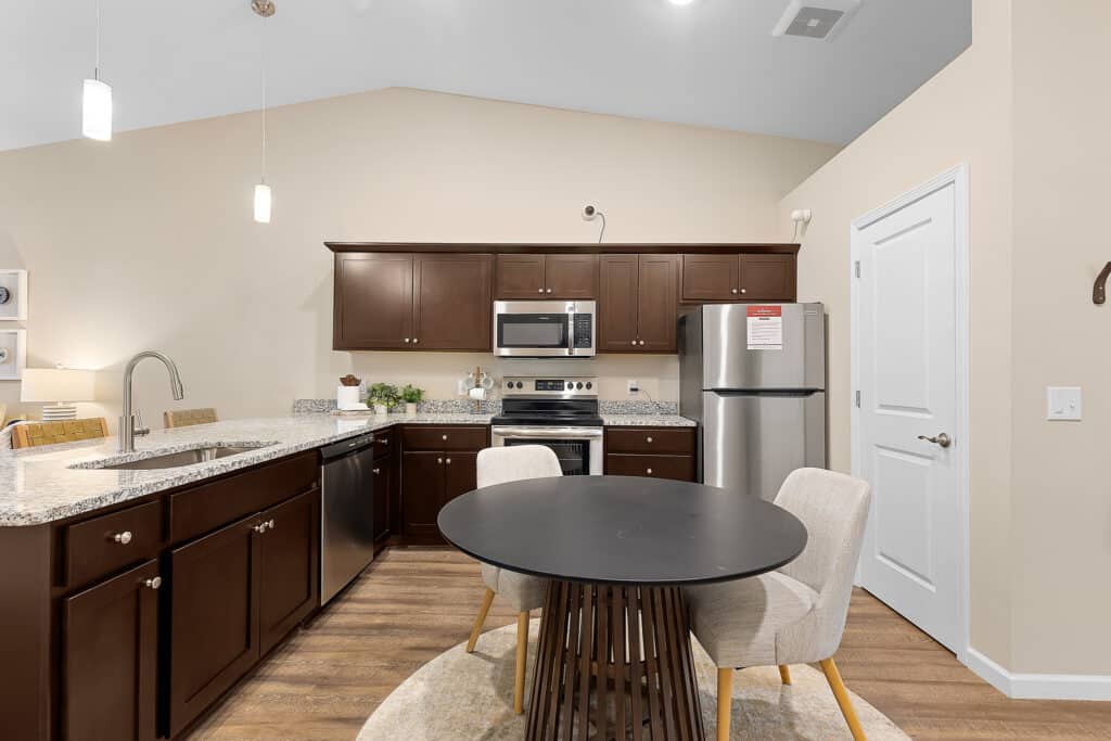 Modern kitchen with dark wood cabinets, stainless steel appliances, a round black dining table, and two beige chairs on a rug. Granite countertops and pendant lighting are visible.