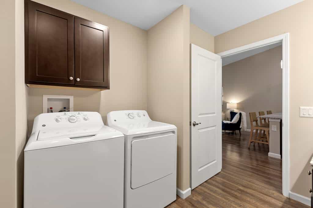 A laundry area with a white washing machine and dryer under dark wooden cabinets, adjacent to an open door leading to a living space with wood flooring.