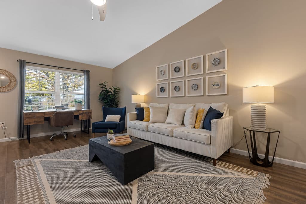 A modern living room with a beige sofa, black coffee table, two lamps, wall art, a desk by the window, wood floors, and neutral-colored decor.