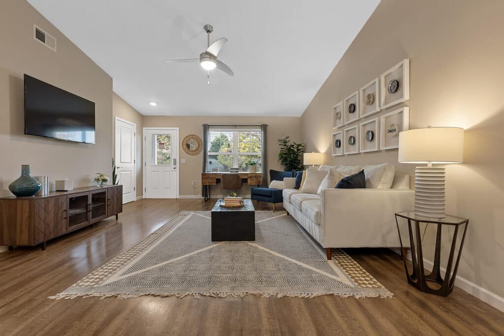 Modern living room with tan walls, wood flooring, a white sofa, wall-mounted TV, and a rug; a desk is placed by the window and decorative art hangs above the couch.