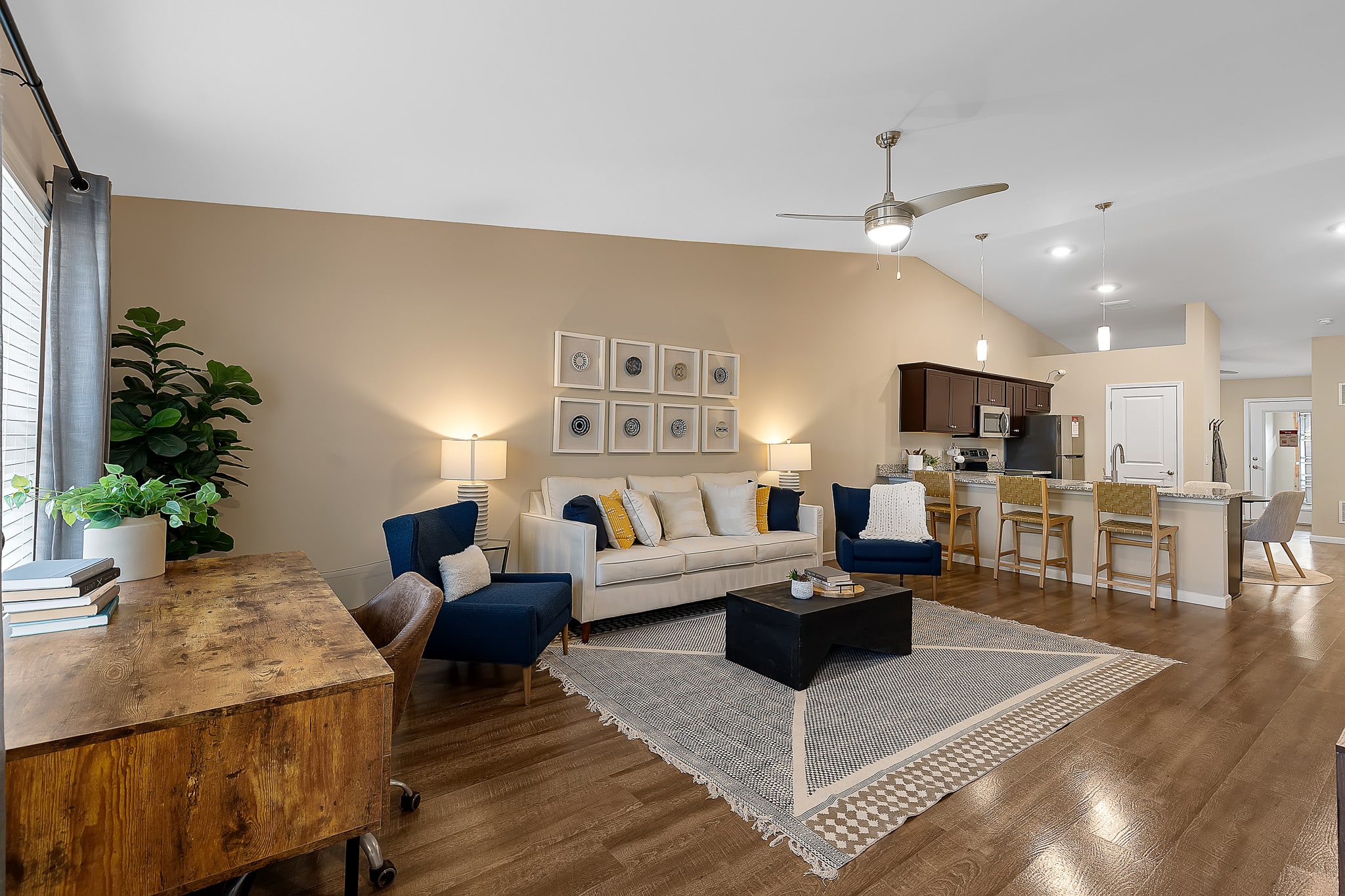 Modern open-plan living room with a beige sofa, blue chairs, a wooden desk, a rug, and a kitchen with bar stools in the background. Light wood flooring throughout.