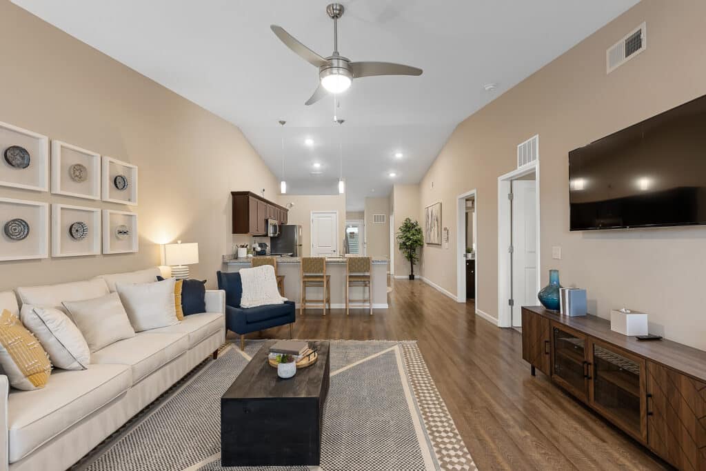 Modern living room with beige walls, a white sofa, blue armchair, wooden coffee table, wall art, TV, and open view into the kitchen and dining area with bar stools.