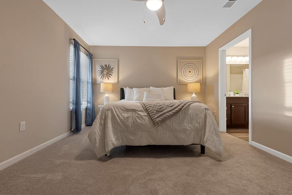 A tidy bedroom with a neatly made bed, two bedside tables with lamps, beige walls, carpeted floor, and an open door leading to a bathroom.