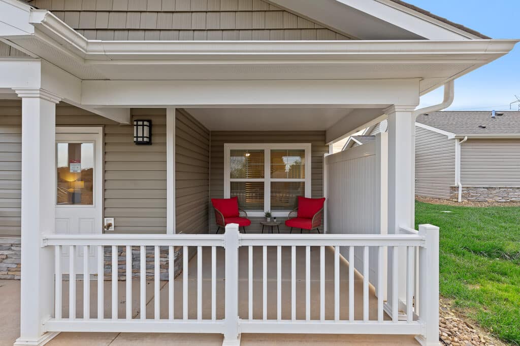 A small covered porch with two red-cushioned chairs and a round table, enclosed by a white railing, attached to a tan-sided house.