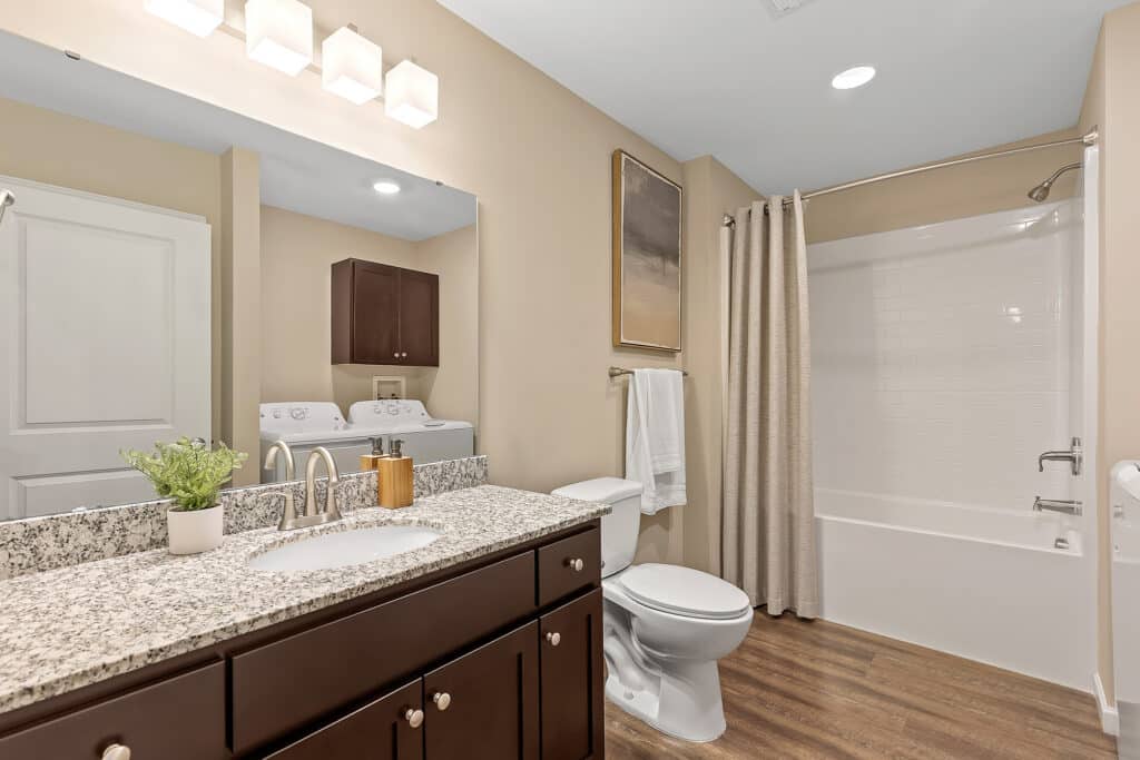 Modern bathroom with granite countertop, dark wood cabinets, white sink, toilet, and bathtub with shower curtain; mirrored wall and washing machine in the background.