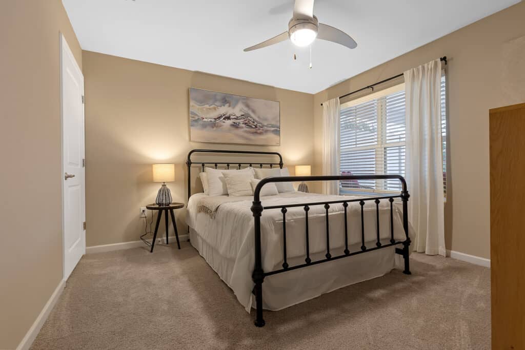 A bedroom with a black metal bed frame, beige walls, carpeted floor, two bedside tables with lamps, a large window with white curtains, and a ceiling fan.