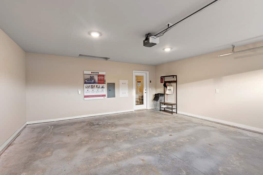 Empty garage with concrete floor, beige walls, ceiling lights, a single door, wall-mounted posters, and a small rack with folded chairs and items on the right.