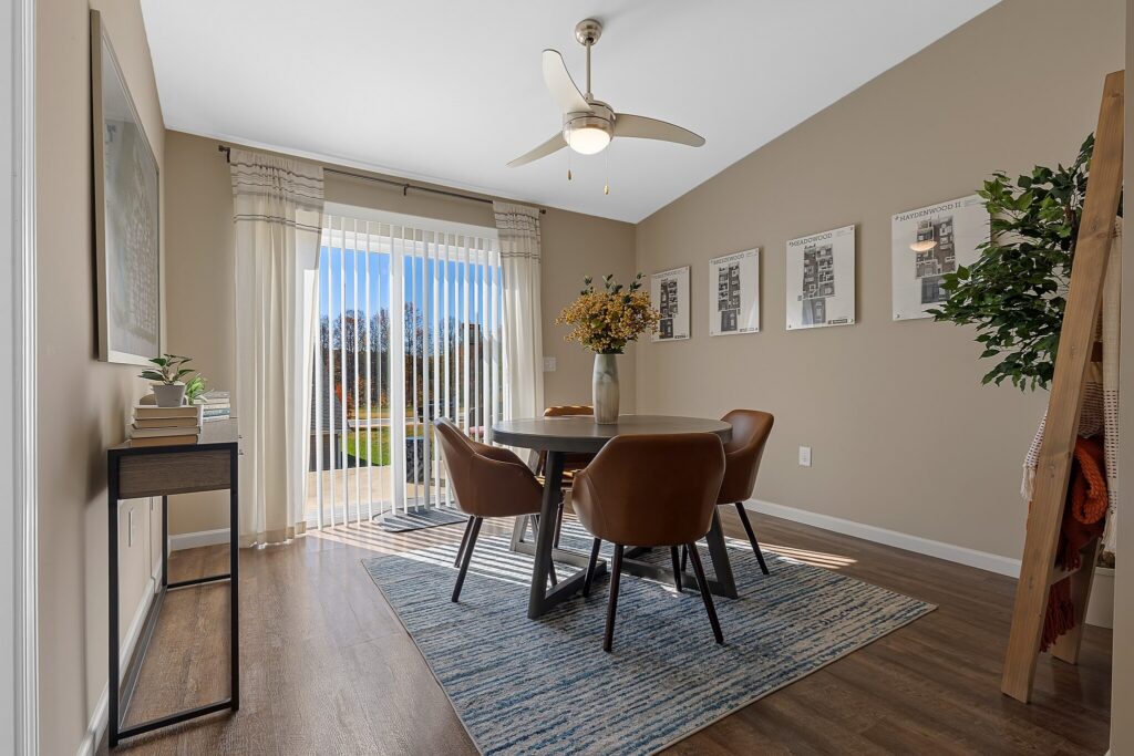 A dining room with a round table, four brown chairs, a rug, a ceiling fan, framed artwork on the wall, and glass doors letting in natural light.