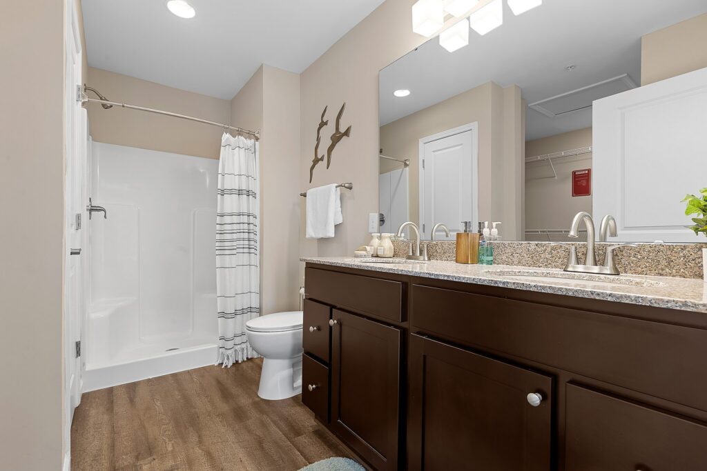 Modern bathroom with a double-sink vanity, granite countertop, large mirror, walk-in shower with striped curtain, toilet, and wood-look flooring.