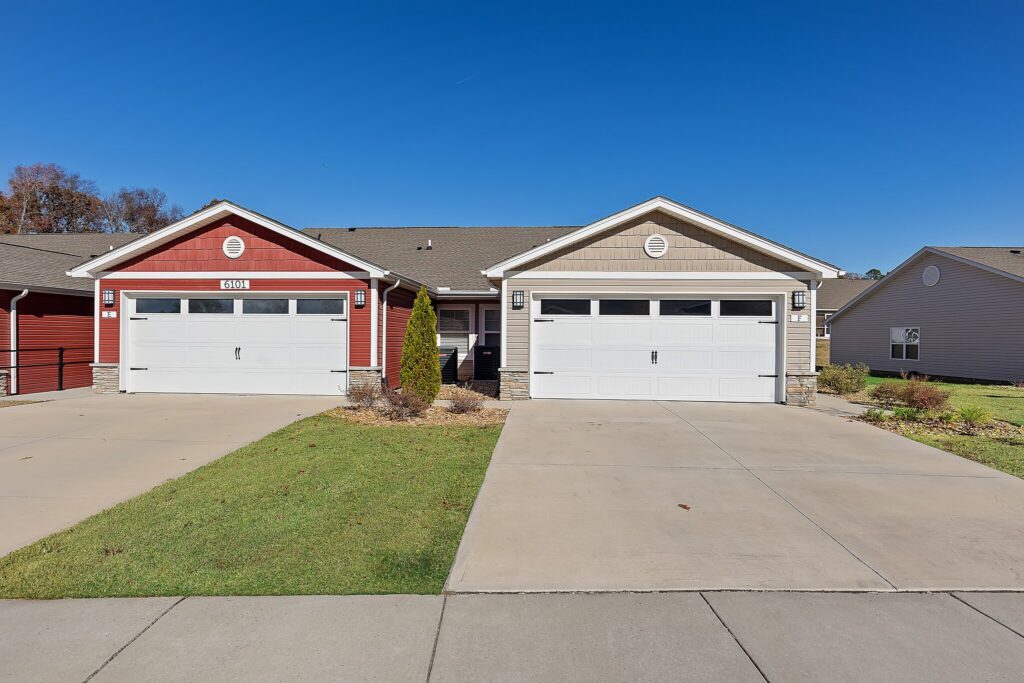 Two attached single-story homes with separate garages, one red and one beige, with well-kept lawns and a clear blue sky.