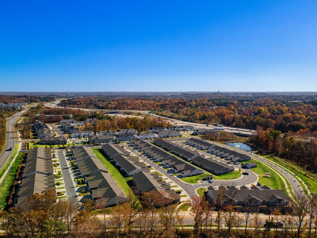 Aerial view of a suburban residential neighborhood with rows of houses, curved streets, and surrounding autumn trees under a clear blue sky.