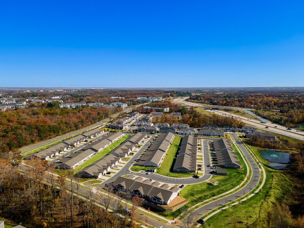 Aerial view of a suburban residential neighborhood with rows of houses, green lawns, curved streets, and nearby highways under a clear blue sky.
