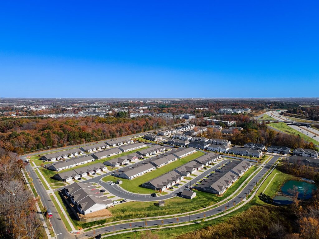 Aerial view of a suburban housing development with rows of identical homes, surrounding roads, nearby trees, and a small pond, under a clear blue sky.