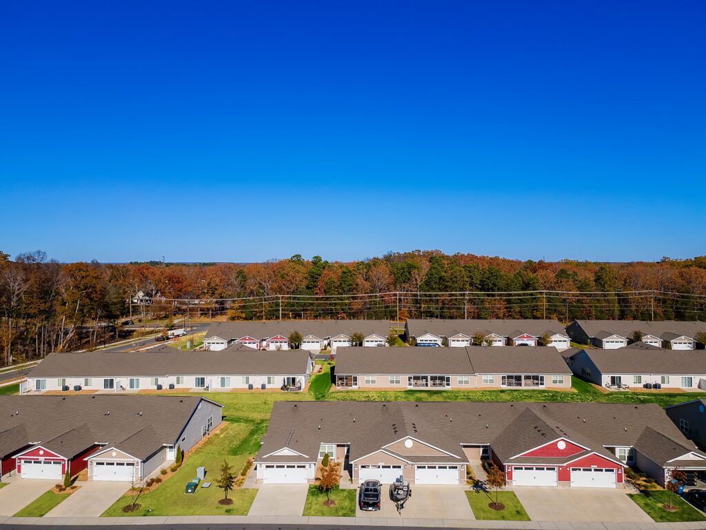 Aerial view of a suburban neighborhood with multiple houses, driveways, cars, and green lawns, bordered by trees under a clear blue sky.