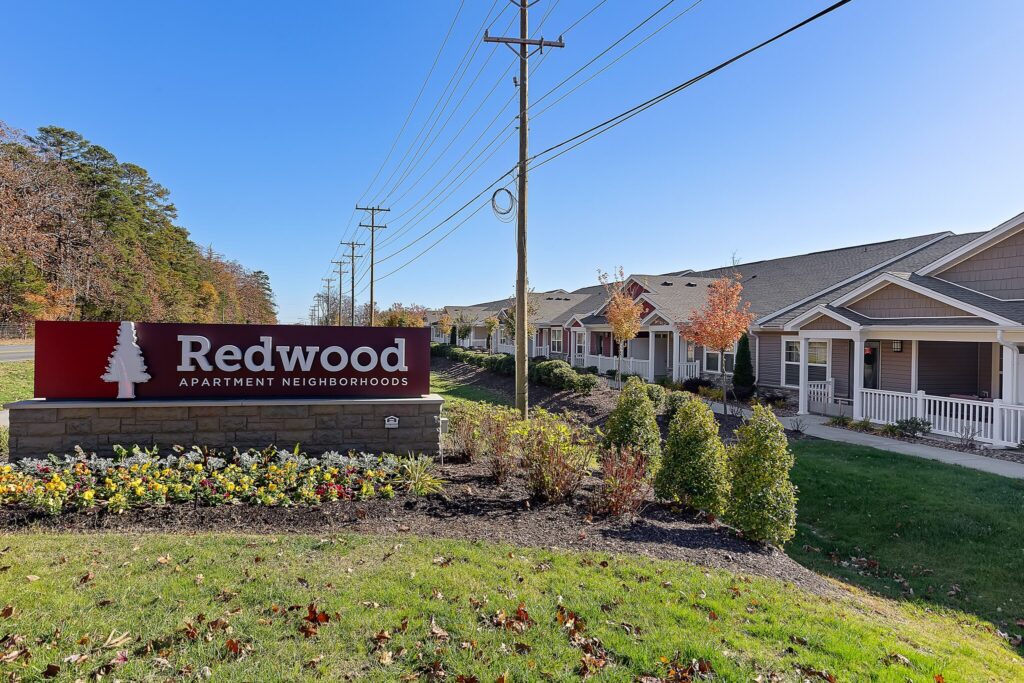 A large red sign reading "Redwood Apartment Neighborhoods" stands in front of single-story apartment buildings on a sunny day.