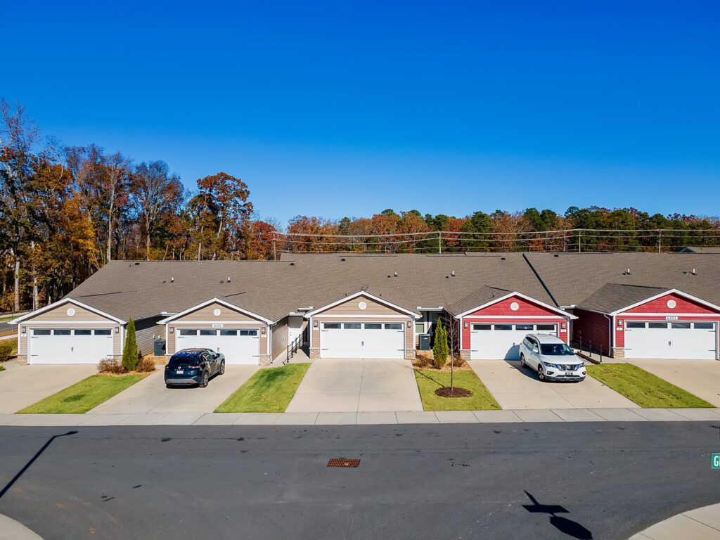 A row of single-story suburban townhomes with attached garages and driveways, two cars parked outside, and autumn trees in the background under a clear blue sky.
