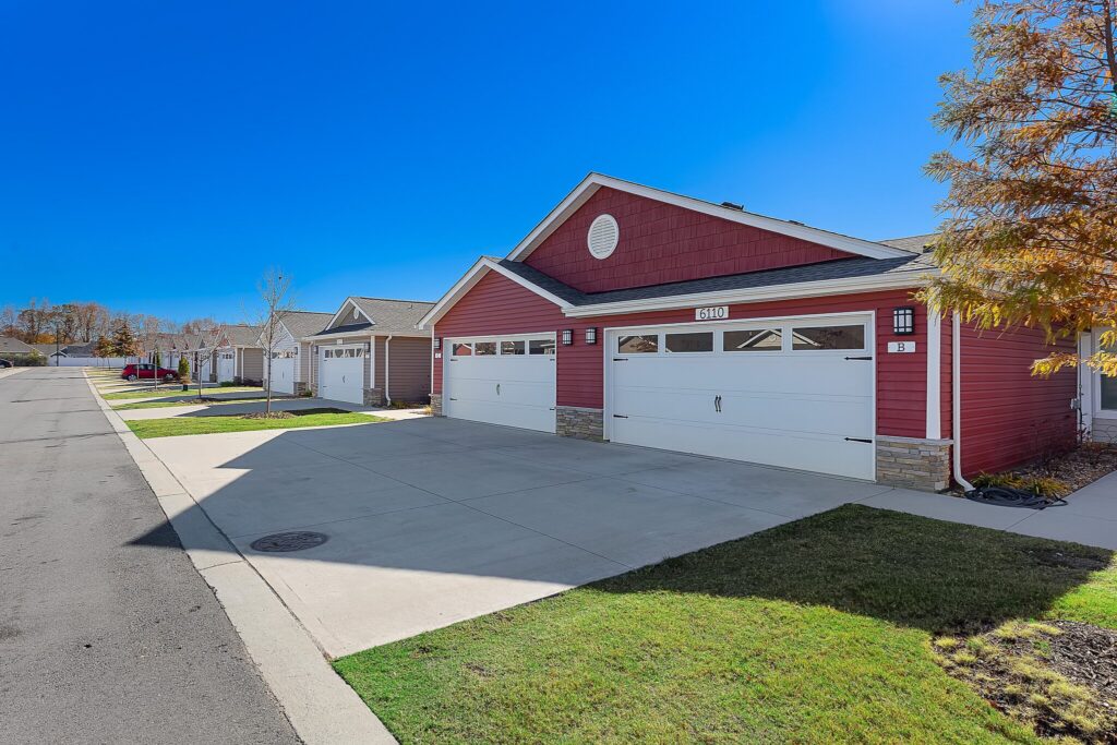 Row of modern, single-story red houses with white garages and driveways on a suburban street under a clear blue sky.
