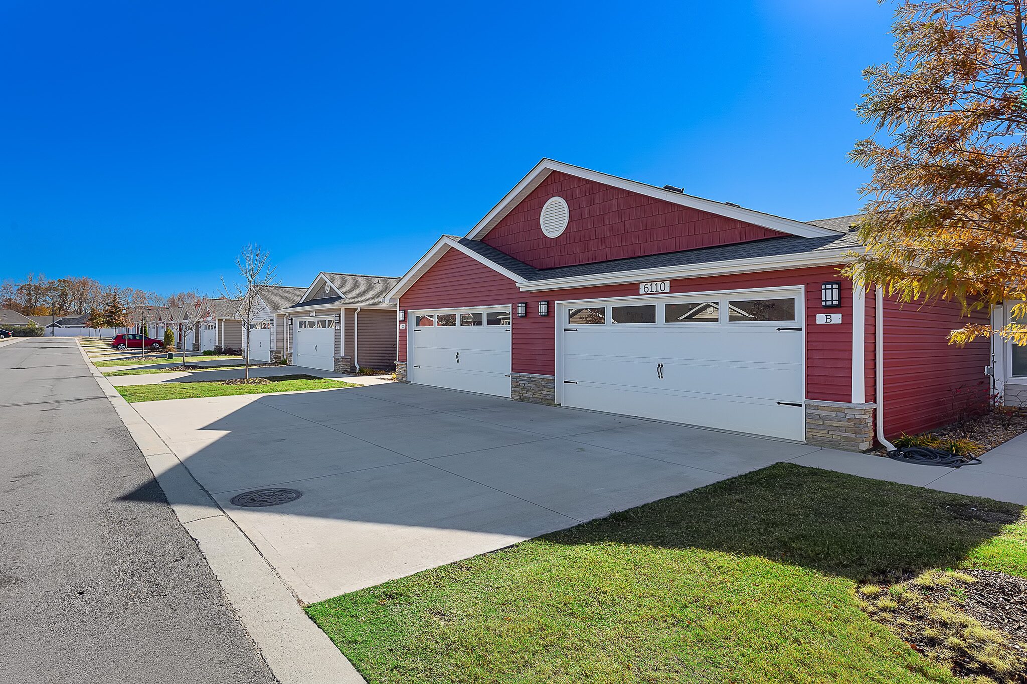 Row of modern, single-story red houses with white garages and driveways on a suburban street under a clear blue sky.
