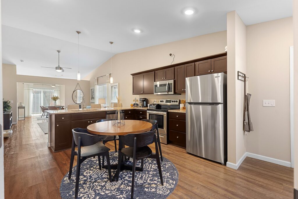 Modern kitchen with dark cabinets, stainless steel appliances, and a round dining table with four chairs on a blue patterned rug, opening into a dining and living area.