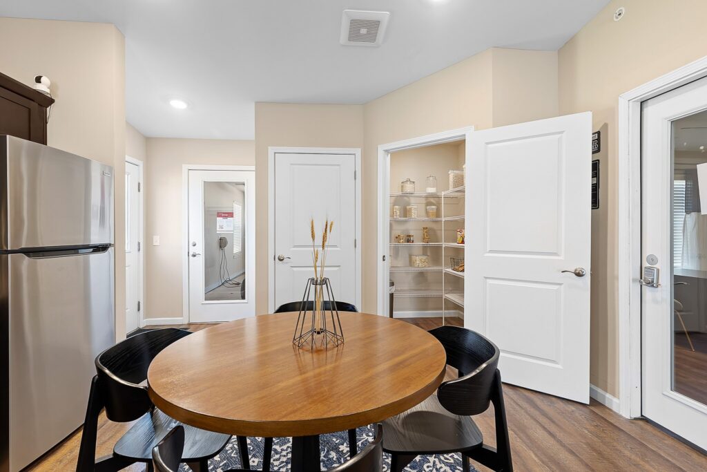 A round wooden dining table with four black chairs is centered in a kitchen area with an open pantry, stainless steel fridge, and doors leading to other rooms.