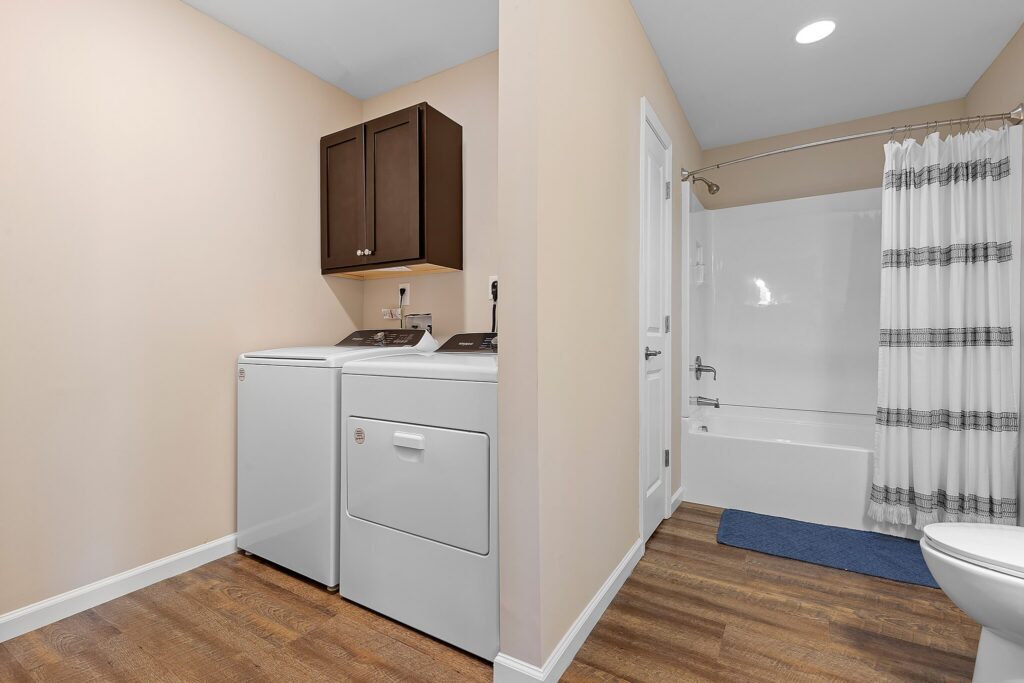 A laundry area with a washer, dryer, and wall cabinets is adjacent to a bathroom with a bathtub, shower curtain, toilet, and blue bath mat on wood-style flooring.