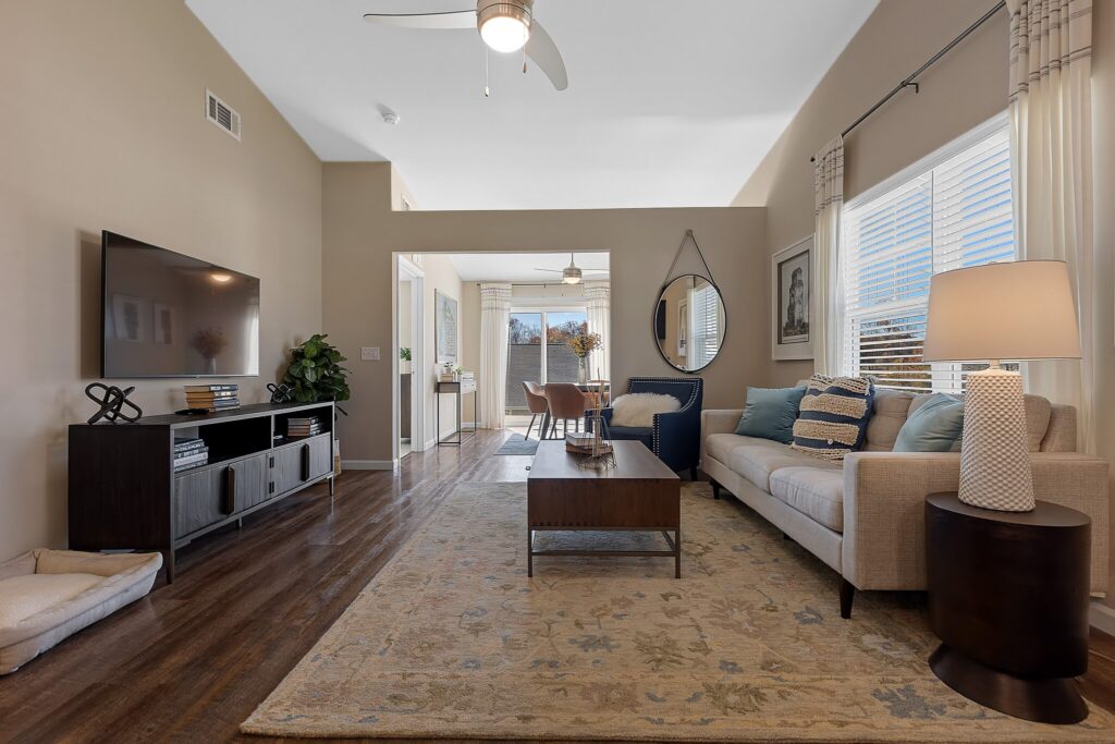 A modern living room with a beige sofa, coffee table, TV, and lamp, featuring wood floors, a large rug, and an adjacent dining area with natural light from windows.