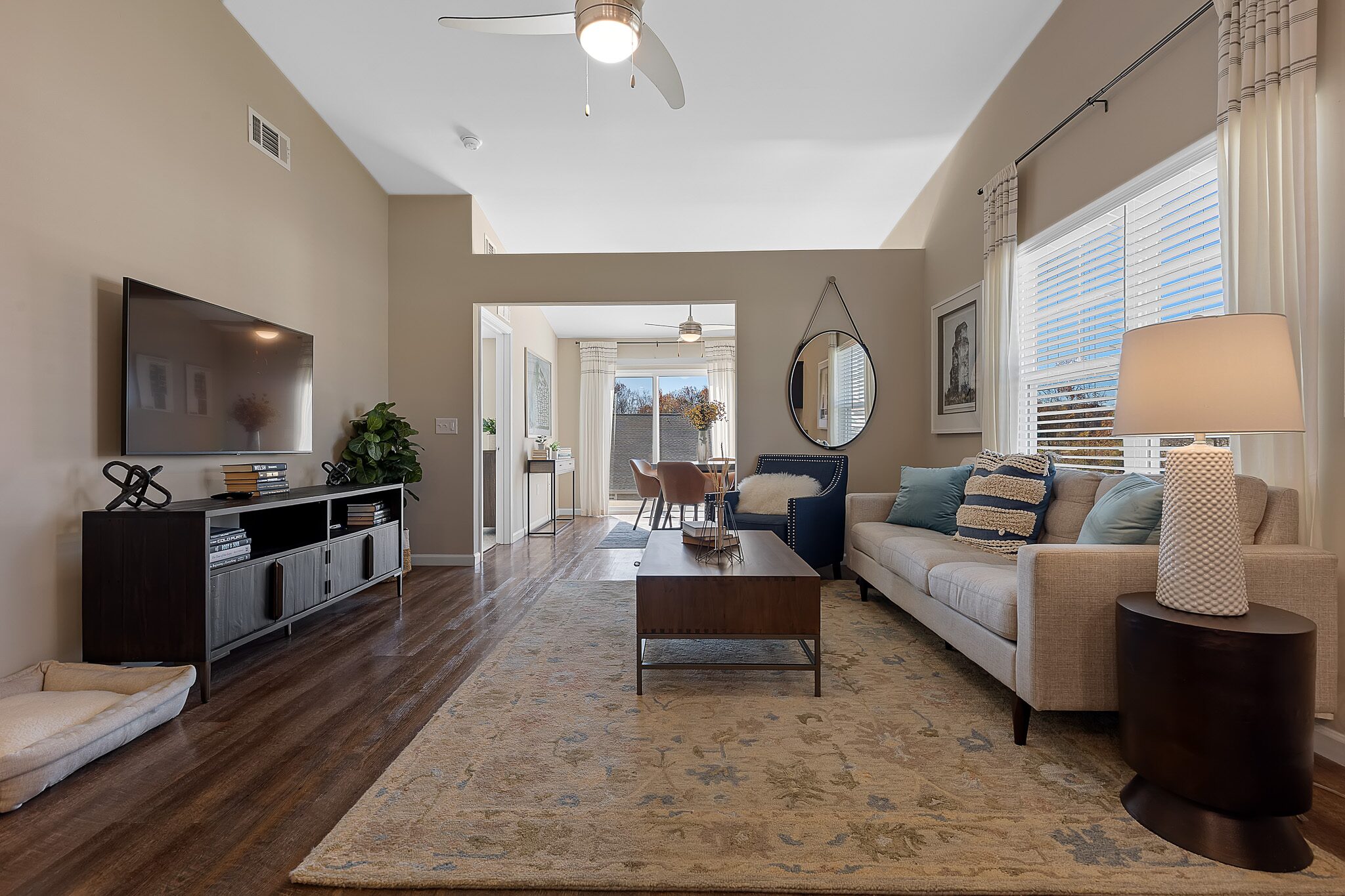 A modern living room with a beige sofa, coffee table, TV, and lamp, featuring wood floors, a large rug, and an adjacent dining area with natural light from windows.