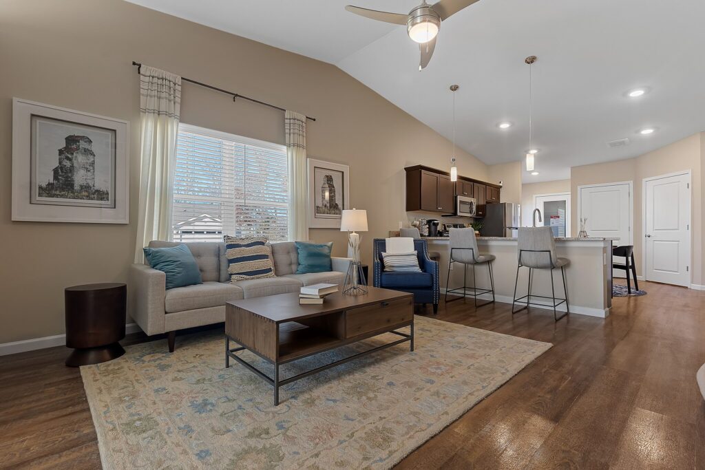 Open-concept living room and kitchen with a sofa, coffee table, armchair, bar stools, and pendant lighting, featuring wood flooring and neutral decor.