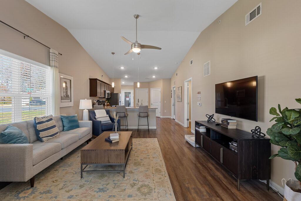Modern living room with beige walls, a gray sofa, a coffee table, a TV on a dark stand, hardwood floors, and an open kitchen in the background. Ceiling fan and large windows are visible.