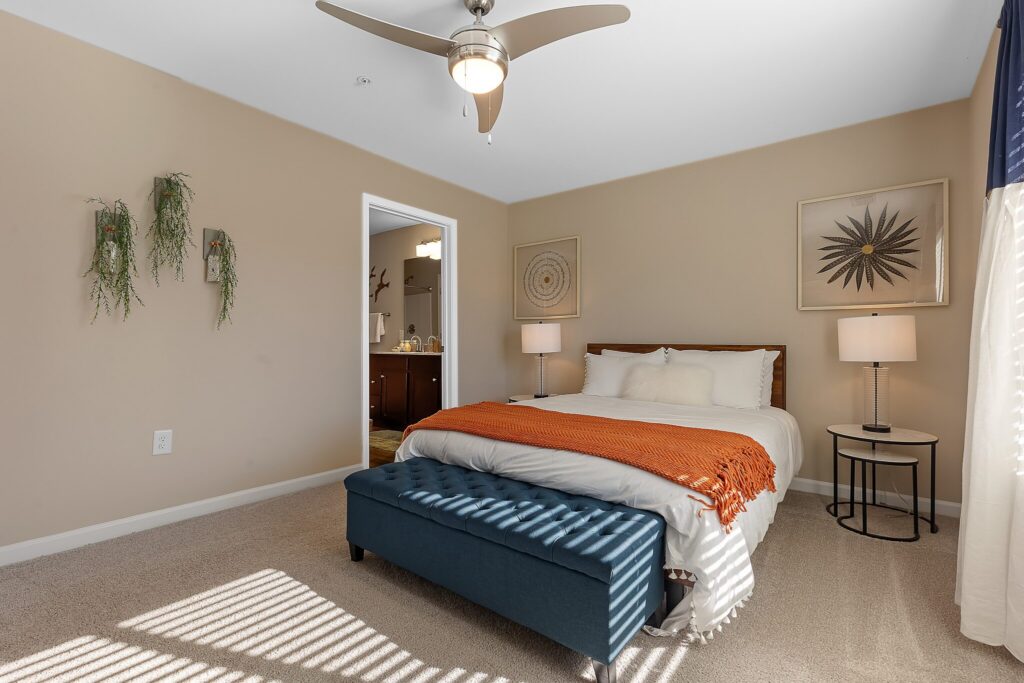A bedroom with a bed, orange throw blanket, blue ottoman, nightstands with lamps, wall art, and an open door leading to a bathroom.