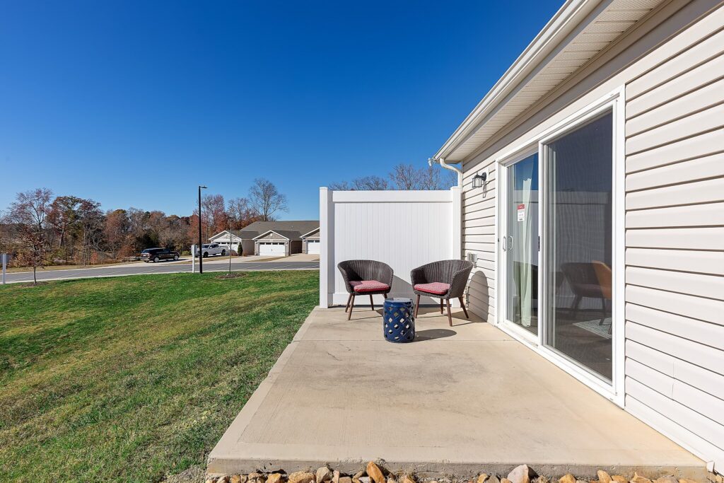 A small concrete patio with two wicker chairs and a side table sits outside a beige house with sliding glass doors and a white privacy fence, overlooking a grassy yard and a residential street.