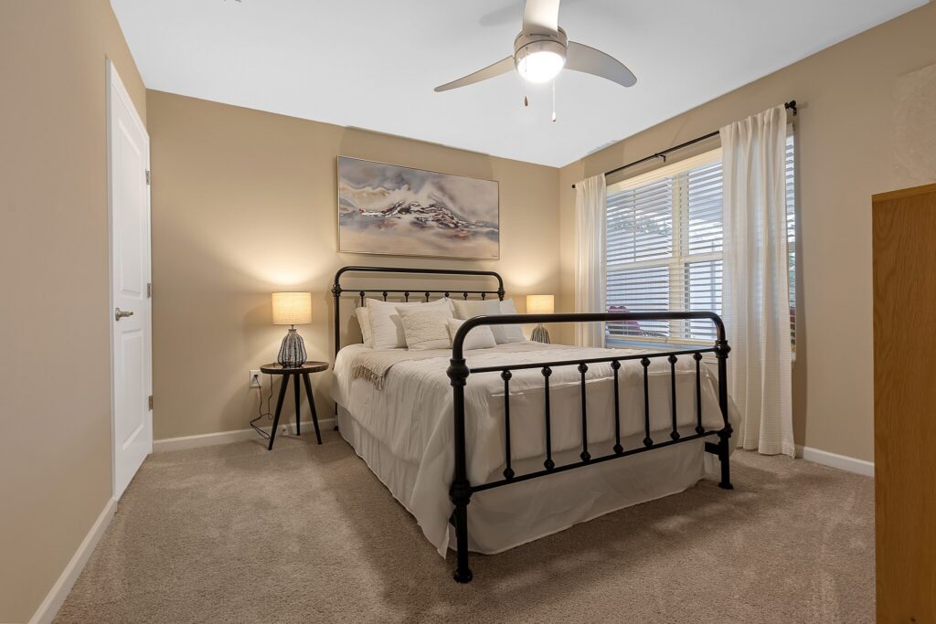A neatly made bed with a black metal frame sits between two nightstands with lamps in a beige-carpeted bedroom with neutral walls, a ceiling fan, and a window with white curtains.