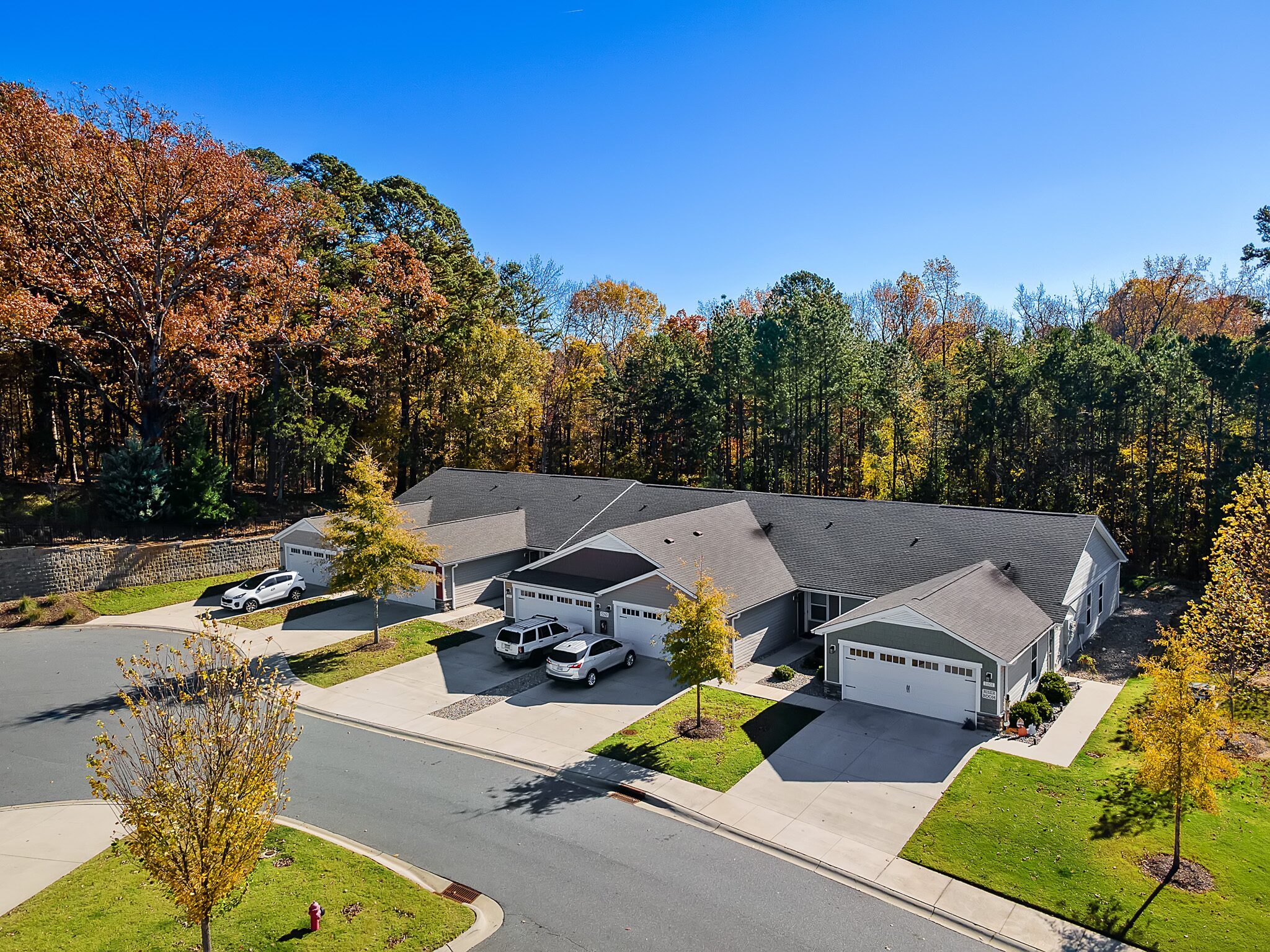 A row of single-story townhomes with attached garages on a quiet cul-de-sac, surrounded by autumn trees under a clear blue sky.
