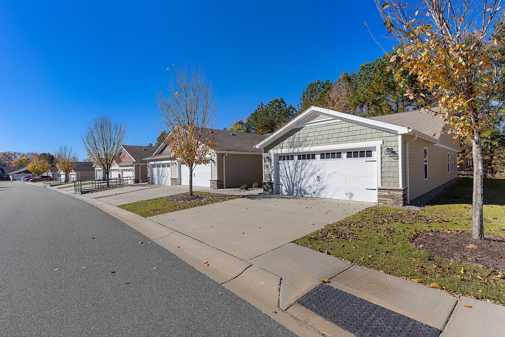 Row of single-story suburban houses with attached garages along a curved residential street, trees with autumn leaves, and clear blue sky.