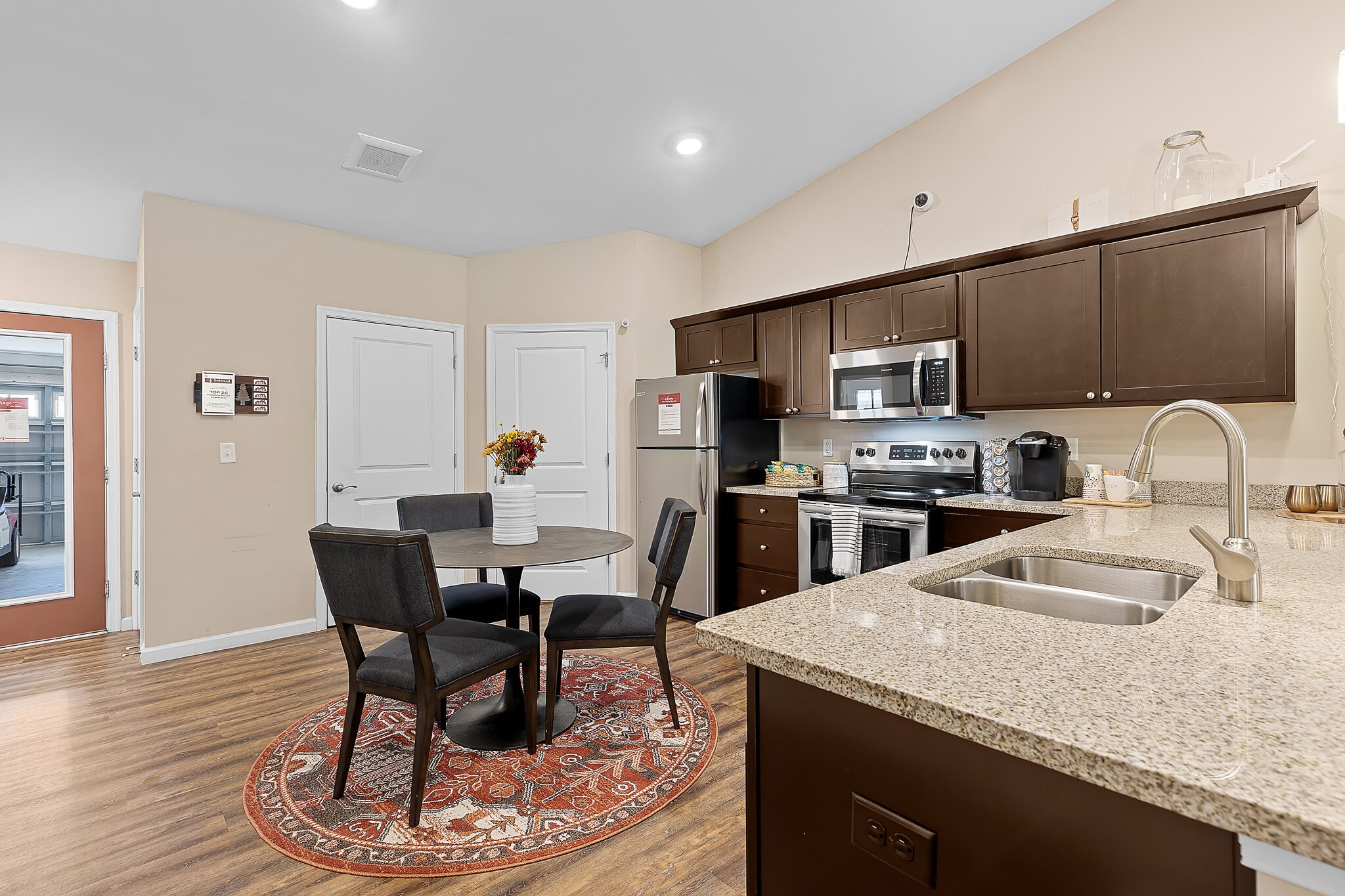 Modern kitchen with dark wood cabinets, stainless steel appliances, a round table with four chairs on a patterned rug, and a vase of flowers as centerpiece.