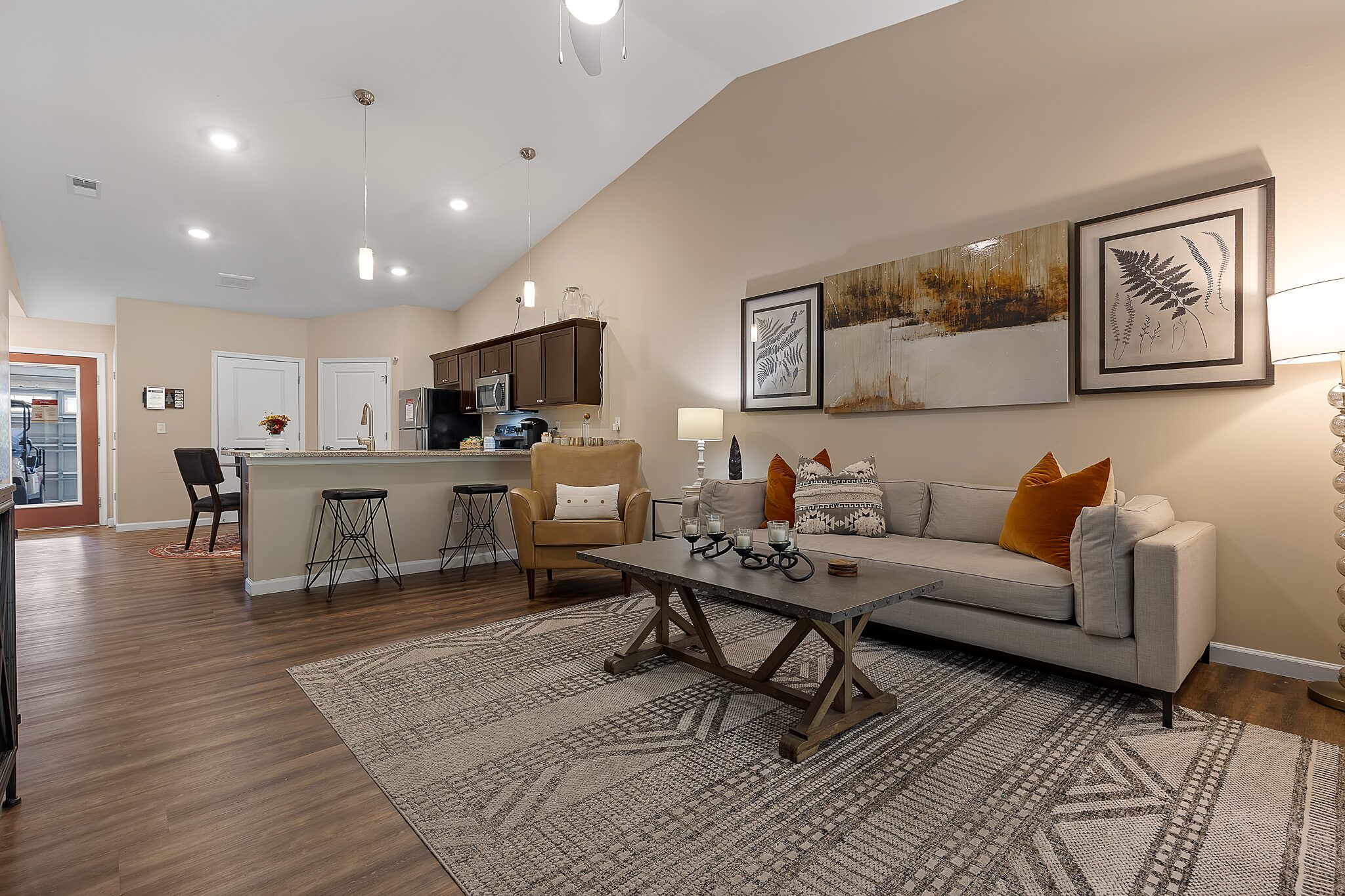 Modern open-concept living room and kitchen with neutral tones, wooden floors, a gray sofa, and bar stools at a kitchen counter under vaulted ceilings.