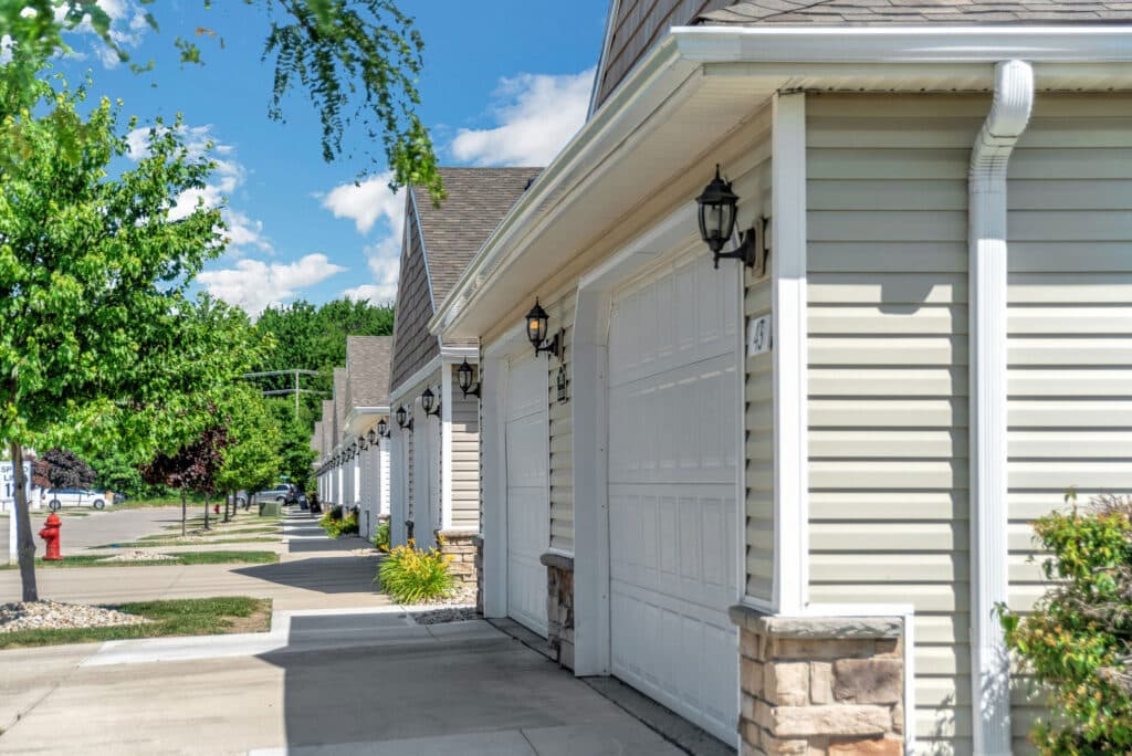 Row of beige garages attached to residential homes, with outdoor lantern lights, small shrubs, and a sidewalk lined with trees under a blue sky.