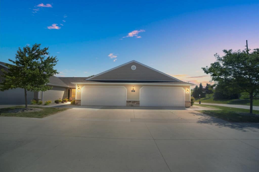 A modern duplex with two white garage doors, illuminated by exterior lights at dusk, with a concrete driveway and trees on either side.