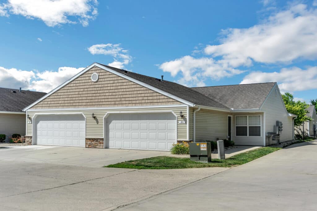 A duplex building with beige siding, two white double garages, a small lawn, and a driveway under a partly cloudy sky.