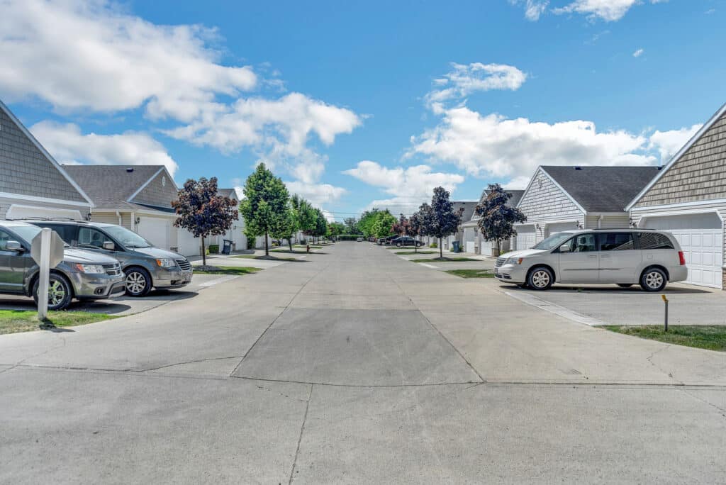 A suburban street with driveways, parked cars, and houses on both sides under a partly cloudy sky.