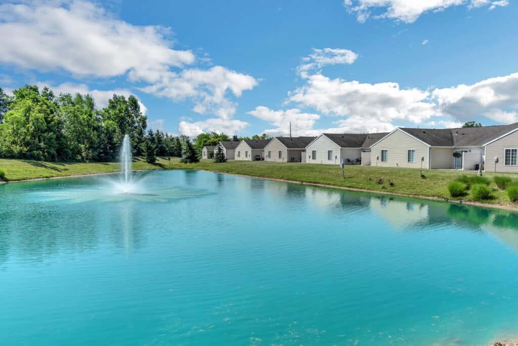 A row of white houses lines a grassy bank beside a pond with a water fountain under a partly cloudy blue sky.